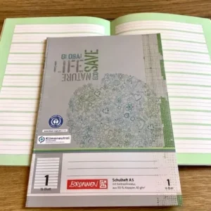 Schulheft A5, aufgeschlagen mit Lineatur 1 vor grünem Kontrasthintergrund, Cover grau mit blaugrüner Weltkugel