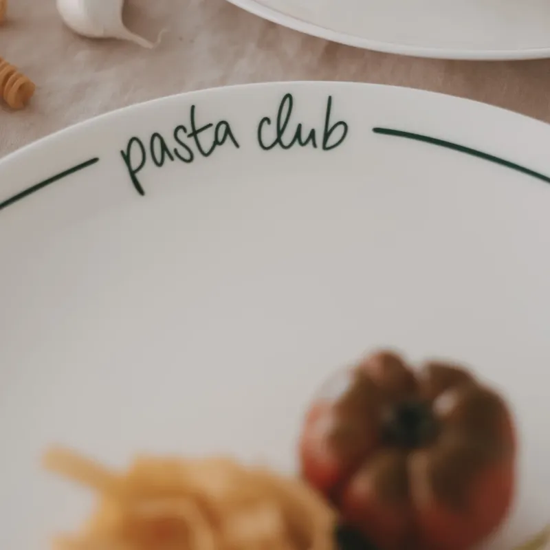 Detailaufnahme des „pasta club“ Schriftzugs auf grün gerandetem Speiseteller mit unscharfer Pasta und Tomate im Vordergrund.