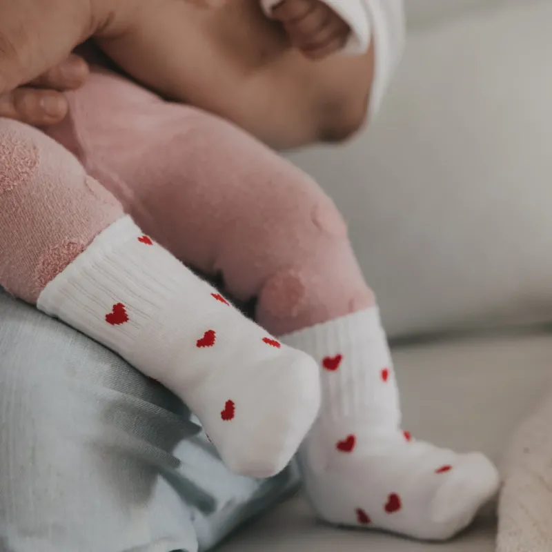 Babybeine mit weißen Herzsocken in Händen gehalten, liebevolle Szene mit weichen Textilien